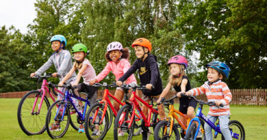 6 children lined up sat on bikes with helmets on. Trees in the background, grass under foot