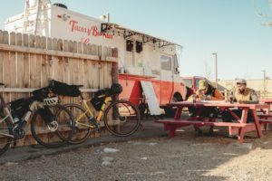2 riders stopped, bikes leaning on a fence, sat on a bench, eating tacos from a taco van
