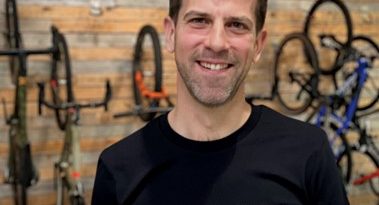 Portrait shot of Remi Rennesson, from the mid torso up, with bikes hung on wood wall under Classified logo behind him