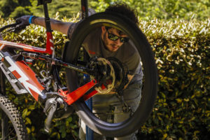 Mechanic with bike in works stand tuning gears, sunglasses on, enjoying the sunshine