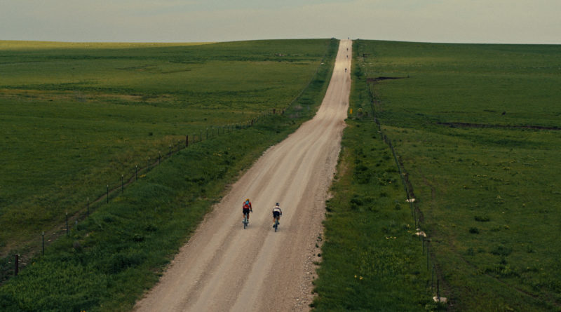Gravel road toward the horizon with lush green grass on either side. Two cyclists riding side by side. What an adventure!