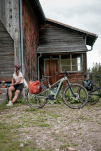 woman sat outside wood cabin with bike sporting packed panniers, tyres muddy, bike clearly being ridden off road