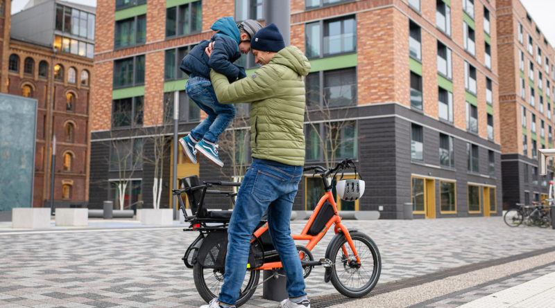 Man lifting child into child seat fitted to the back of the Tern Short Hall, with the backdrop being a n urban setting, possibly an apartment block