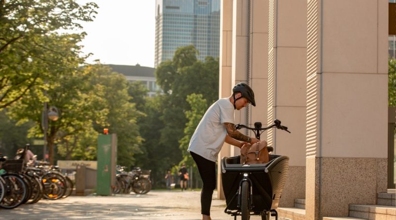 Man loading bag into cargo bike on a sunny urban sidewalk with trees to to the foreground and highrise buildings in the background