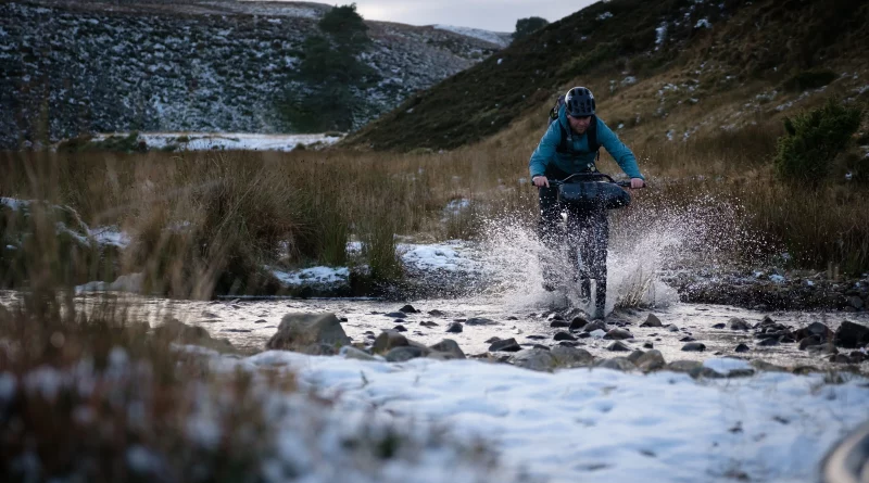 MTB fitted with bikepacking bar bag, being ridden through a stream in a gully, with light snow on lowland grounds
