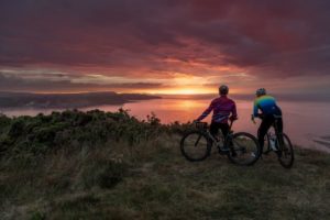 2 riders at a clif top looking out at a stunning pink, orange, sun set with cloud form on the horizon