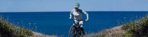 Woman riding mountain bike up over crest with sea as backdrop 