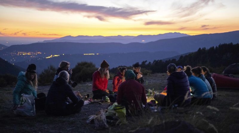 Women sat around a camp fire on a beautiful sun set evening, just as dusk begins to fade, mountains on the skyline in the background