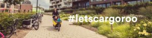 #letscargoroo banner with woman riding cargo bike toward camera in green urban space