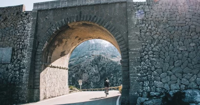 Old stone bridge with road winding through, cyclist riding into the sunlight