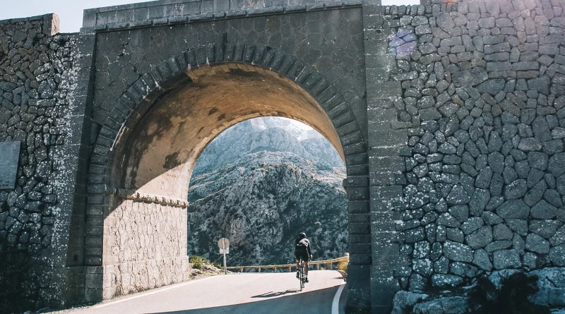 Old stone bridge with road winding through, cyclist riding into the sunlight