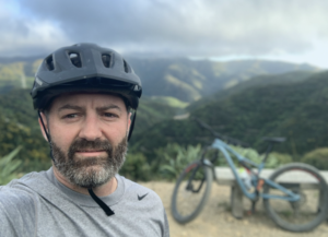 Adam Barnes, Bike Matric Tech Co-Founder, paused on a Mountain bike ride, with green hills behind him and bike to one side