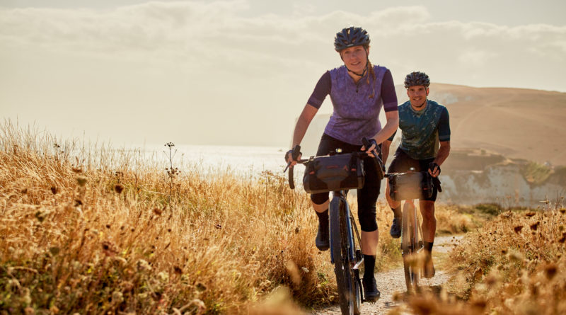 Woman and man riding gravel bikes on cliff top coastal path with burnt orange grass and sea views behind them