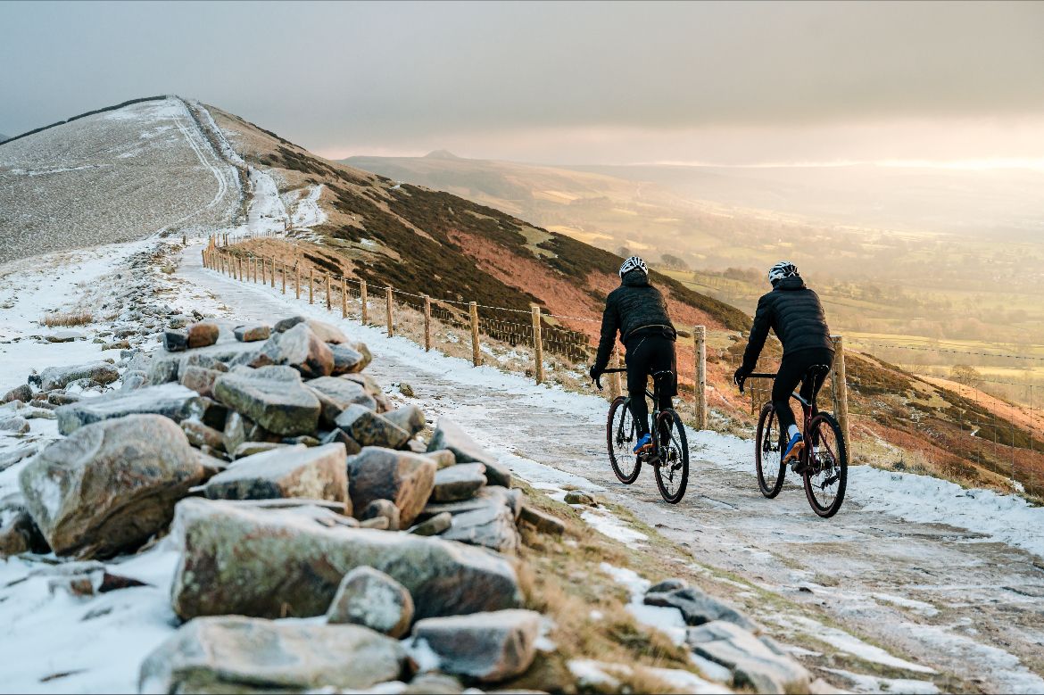 2 Riders on Gravel bikes pedalling along a hill top path covered in light snow with winter sun shining on the right of the image