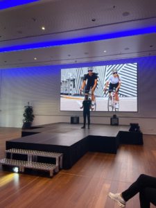 Assos presenter on stage at SS24 launch event with image of man and women on bikes in Assos kit behind him