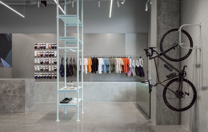 Long shot of MAAP LaB with marble floor, light grey walls, and cleanly merchandised socks and jerseys in the background. A bike handing from its front wheel in the mid foreground