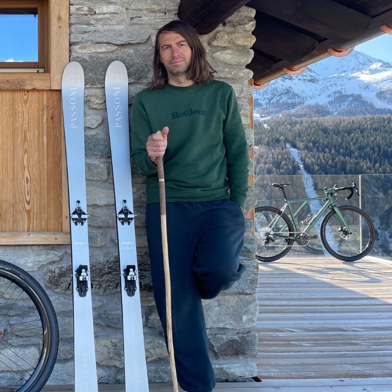 Matteo Cassina, owner of Gruppo Media, leaning against stone wall ski chalet with Passoni branded skis lent against wall and 2 bikes in shot 