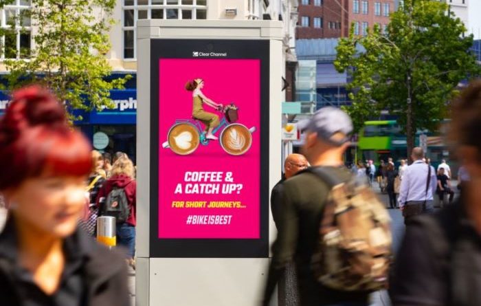 Digital billboard on highstreet with people walking up and down. 'Coffee and a catch up? slogan with digital cartoon of person riding bike with late art wheels