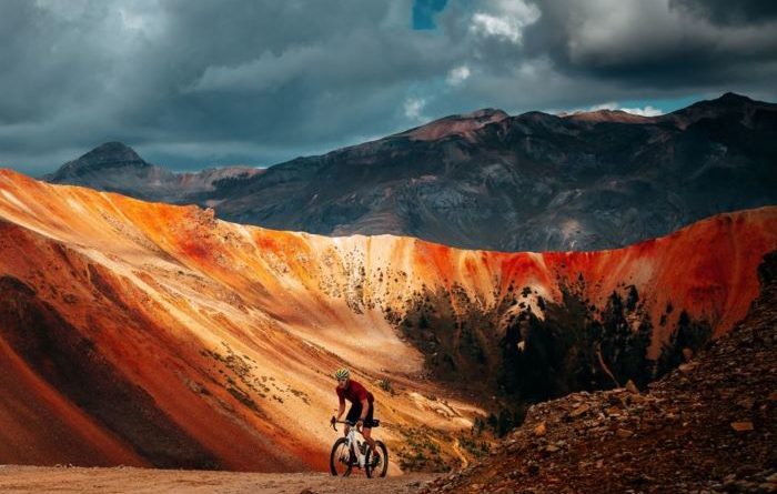 Gravel rider making way out of bowl like red earthen feature with mountains in backdrop