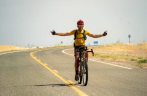 Ulrich Bartholmoes - Tour Divide 2023 - riding arms outstretched on an open road with dust and burnt grass stubs either side
