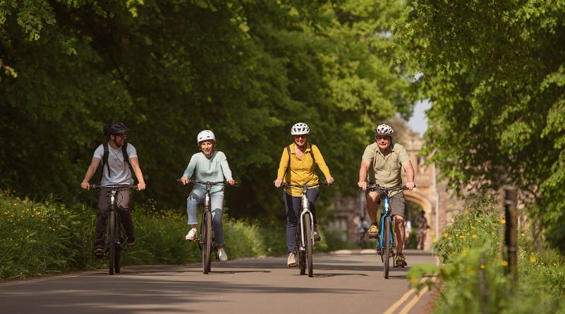 4 riders, men and women spanning age ranges, pedalling bikes along side by side on a cycle path with trees on either side